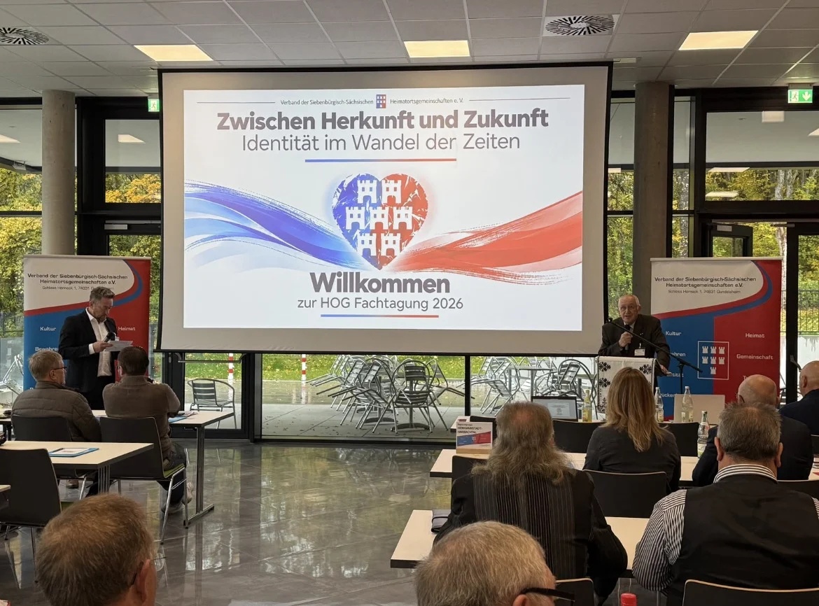 Begrüßung zur HOG-Fachtagung 2026: Leinwand mit dem Motto „Zwischen Herkunft und Zukunft – Identität im Wandel der Zeiten“, Redner am Pult und Moderator, davor die Teilnehmer an Tischen