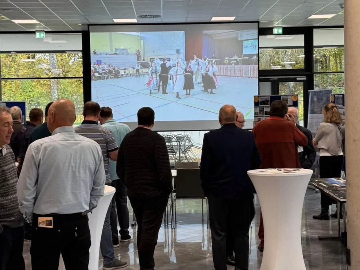 Teilnehmer der HOG-Fachtagung im Gespräch an Stehtischen, auf der Leinwand ein Video mit Tanzgruppen in siebenbürgisch-sächsischer Tracht